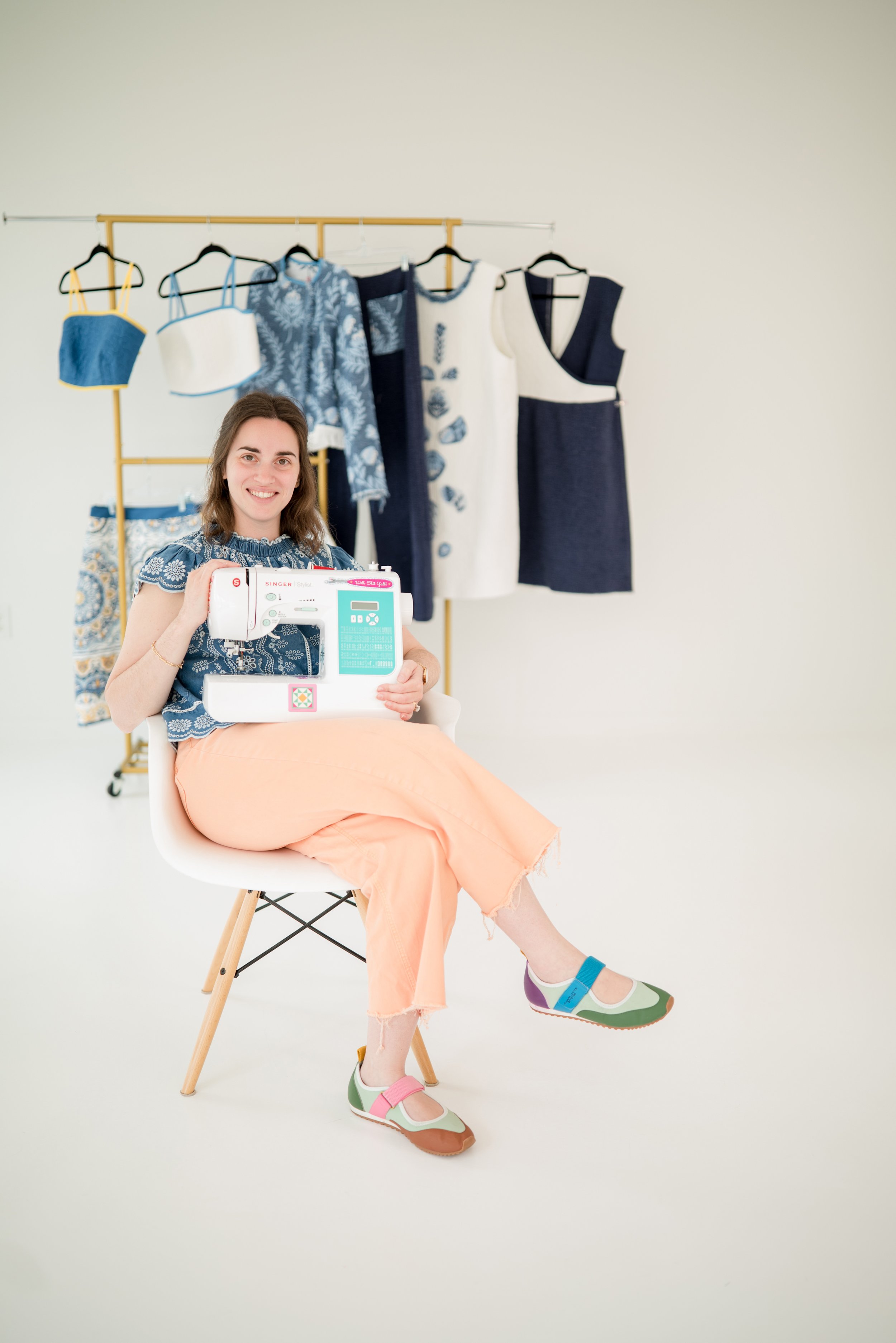 Brigitte of Speedy B Stitchery seated at her sewing machine in front of a rail of handmade garments